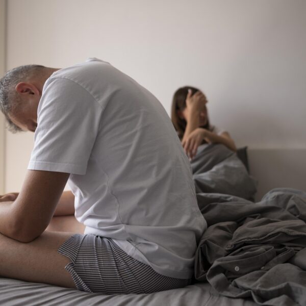 Hombre sentado al borde de la cama mientras una mujer permanece detrás de él. Título: Cambios en el deseo sexual