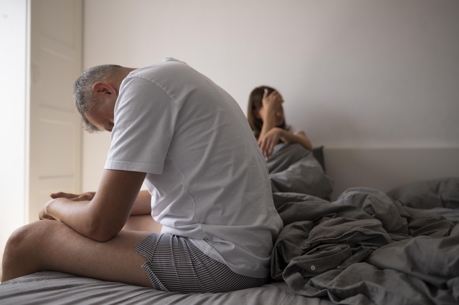 Hombre sentado al borde de la cama mientras una mujer permanece detrás de él. Título: Cambios en el deseo sexual