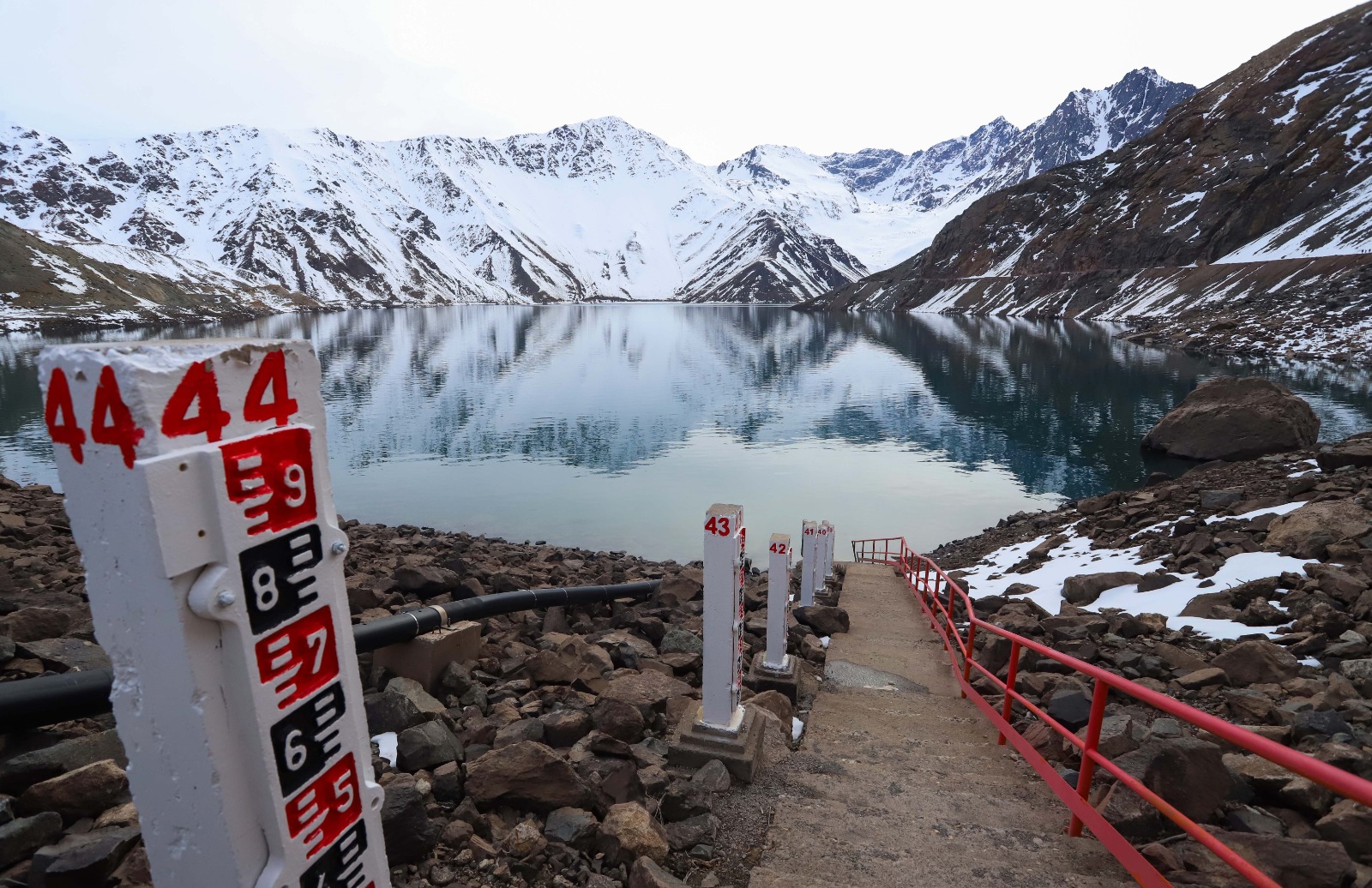 Embalse rodeado de montañas nevadas con reglas para medir el nivel del agua en primer plano.