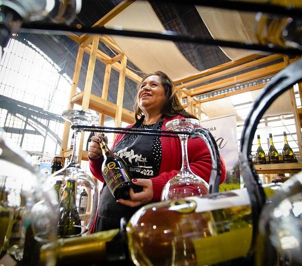 Productora sostiene una botella de vino entre copas y botellas exhibidas en un puesto.