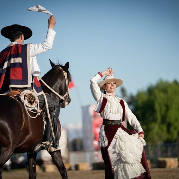 Jinete a caballo y bailarina con vestimenta tradicional chilena levantan pañuelos durante una presentación ecuestre.