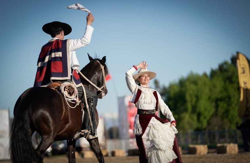 Jinete a caballo y bailarina con vestimenta tradicional chilena levantan pañuelos durante una presentación ecuestre.
