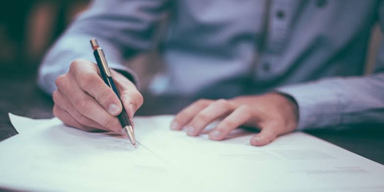 Persona firma un documento con un lápiz sobre una mesa.