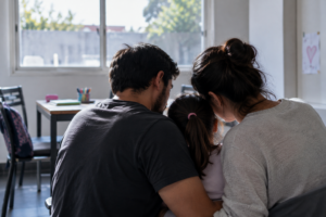 Dos adultos acompañan a una niña en una habitación iluminada por luz natural.